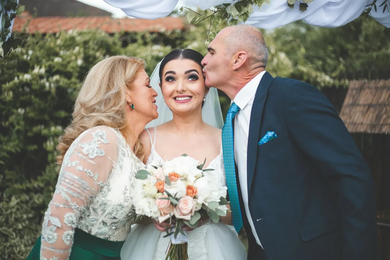 A bride smiling up while her mother and father kiss her cheeks on her wedding day.
