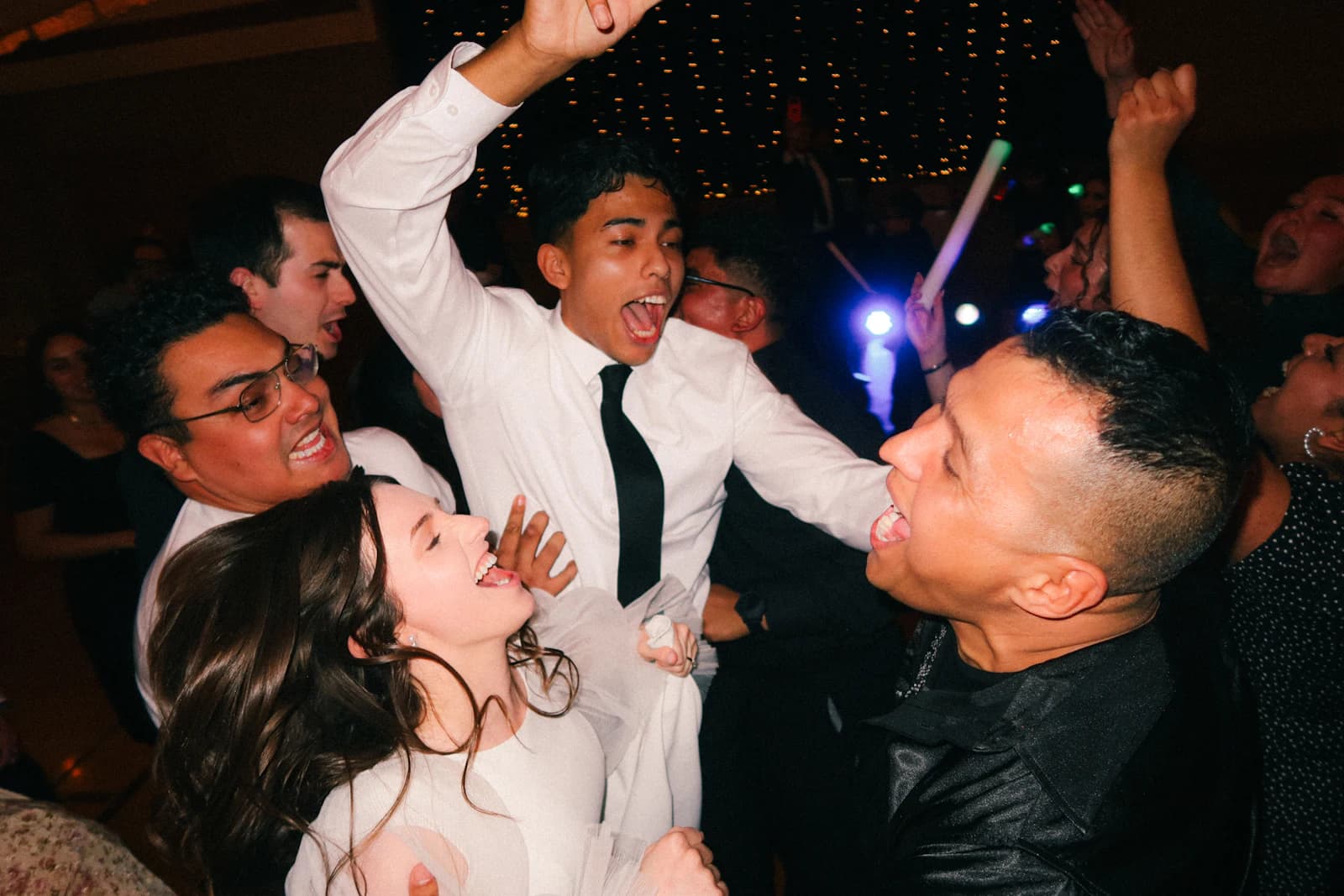 A packed dance floor under warm string lights, guests mid-laugh with their arms up.