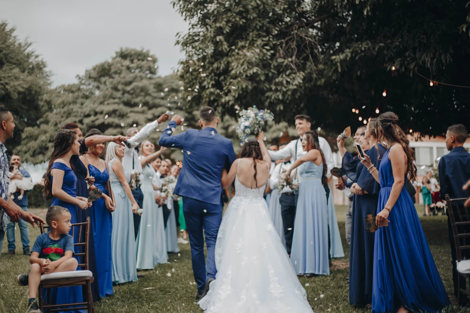 A couple walking back down the aisle outdoors as the wedding party throws petals over them, guests laughing on both sides.